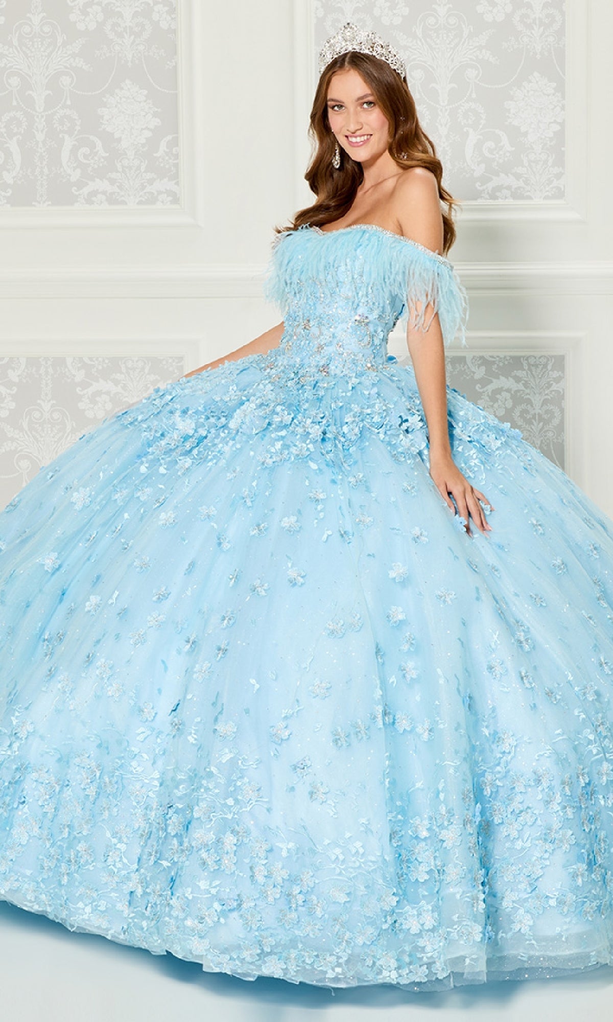 A woman wearing the Quinceanera Dress PR30115 By Princesa—a light blue, off-the-shoulder gown with floral details and a tiara—stands in front of an ornate white wall, smiling at the camera.